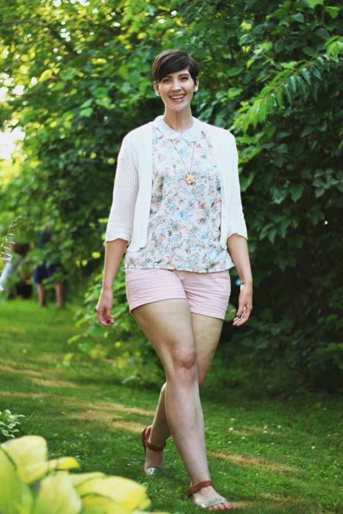 hannah is wearing a pastel floral peter pan collar blouse, a short white cardigan, red and white striped shorts, and glittery sandals. her hair is styled in a short pixie cut. she's standing in greenery.