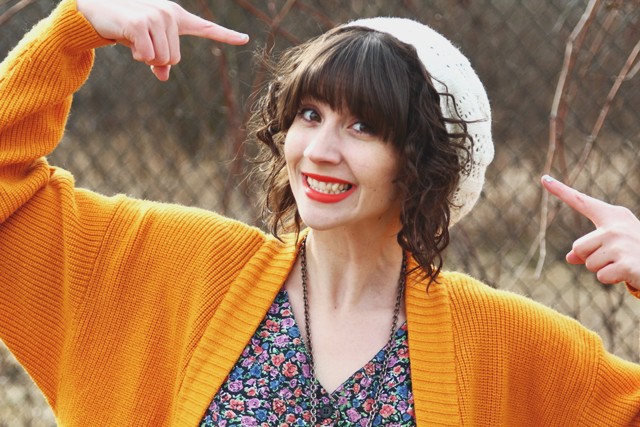 Curly brown hair and a white slouchy knit hat. Bright outfit with orange lipstick.