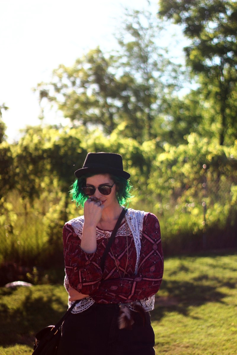 white female standing in a sunny, grassy field. wearing boho style with a red peasant blouse, black pork pie hat, black jean shorts, and white lace vest.