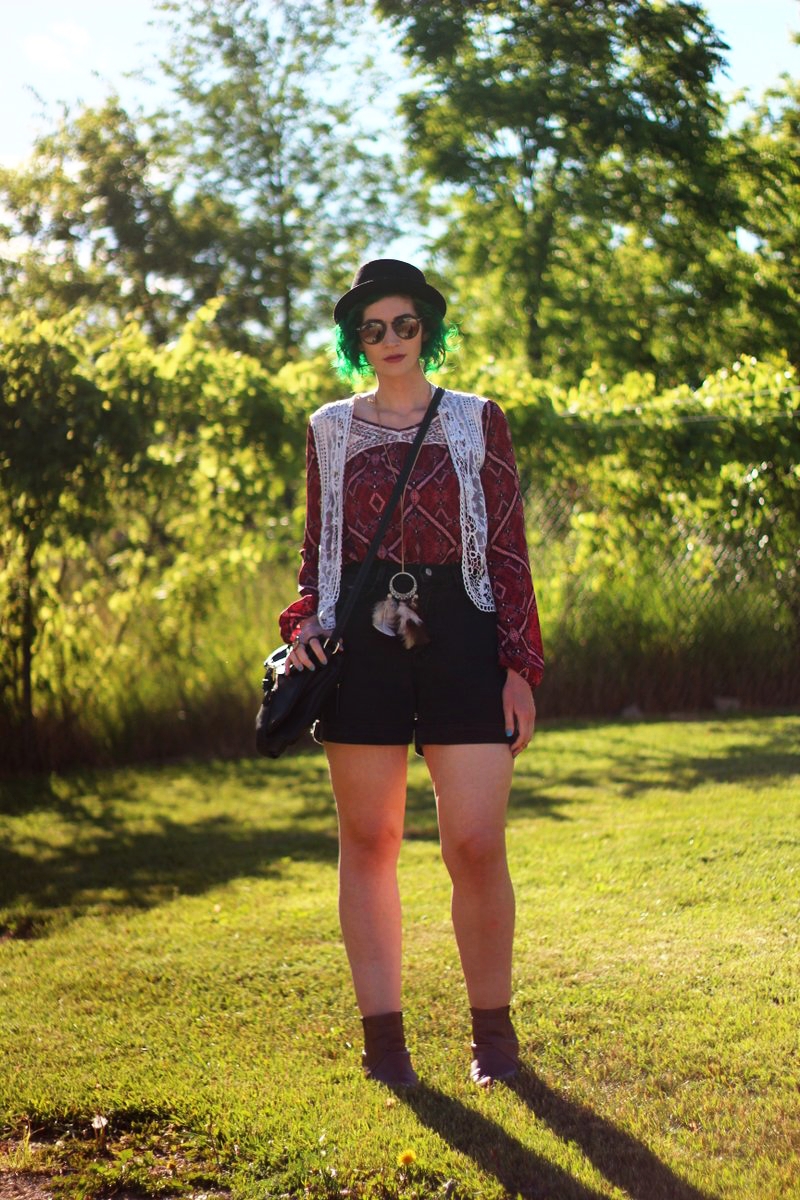 white female standing in a sunny, grassy field. wearing boho style with a red peasant blouse, black pork pie hat, black jean shorts, vest, purse, and boots.