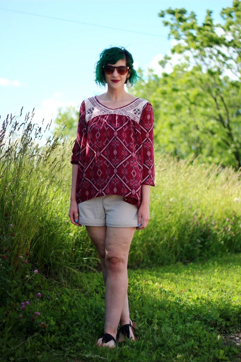 white female standing in a sunny, grassy field. wearing a boho style red peasant blouse, beige striped shorts, and sandals.