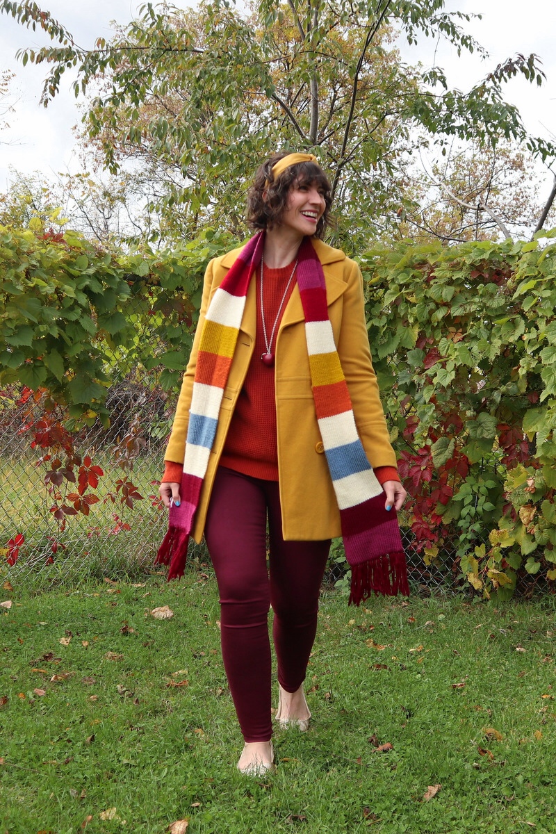 Hannah has short shaggy curls adorned with a yellow headband, in front of a leafy background, wearing an orange sweater, yellow jacket, striped scarf, and burgundy jeggings