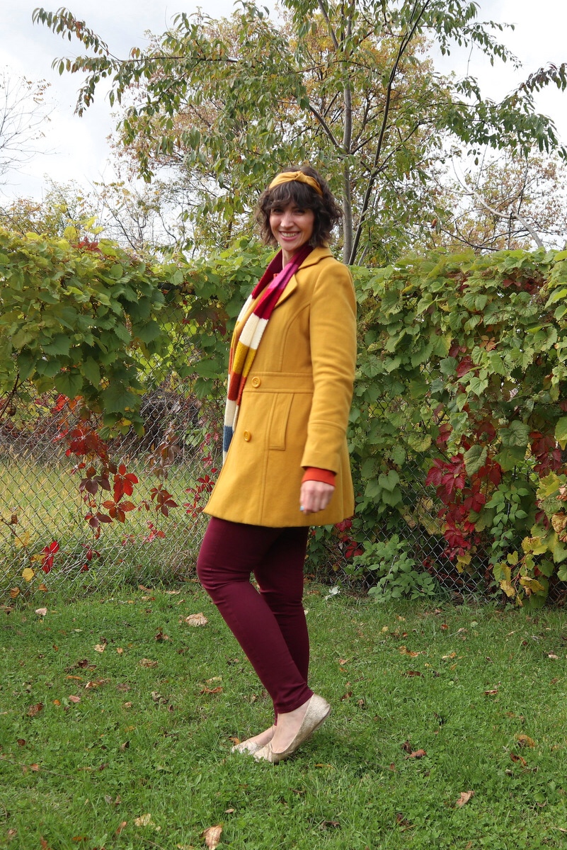 Hannah has short shaggy curls adorned with a yellow headband, in front of a leafy background, wearing an orange sweater, yellow jacket, striped scarf, and burgundy jeggings