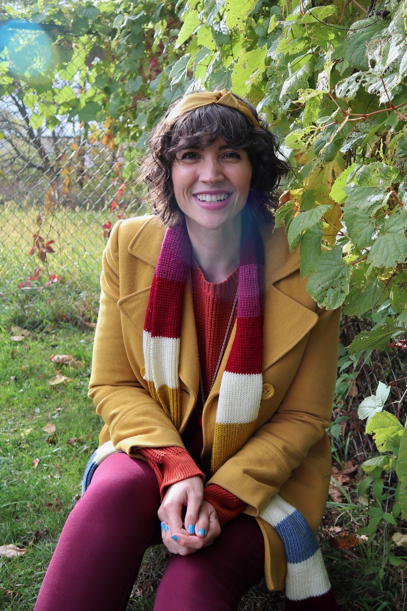 Hannah has short shaggy curls adorned with a yellow headband, in front of a leafy background, wearing an orange sweater, yellow jacket, and striped scarf