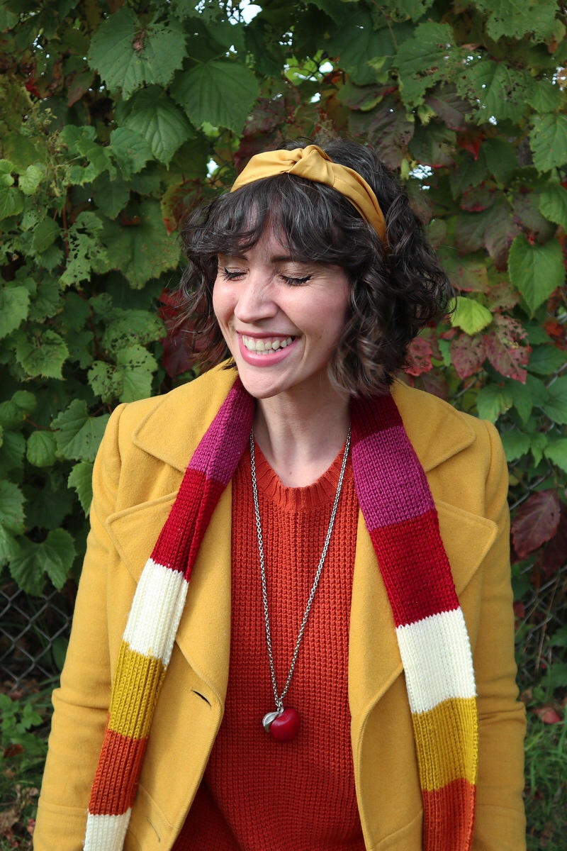 Hannah has short shaggy curls adorned with a yellow headband, in front of a leafy background, wearing an orange sweater, yellow jacket, and striped scarf