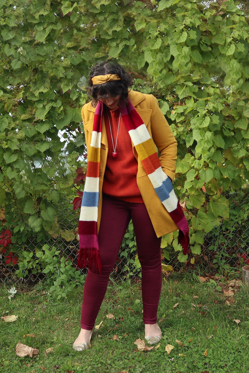 Hannah has short shaggy curls adorned with a yellow headband, in front of a leafy background, wearing an orange sweater, yellow jacket, striped scarf, and burgundy jeggings