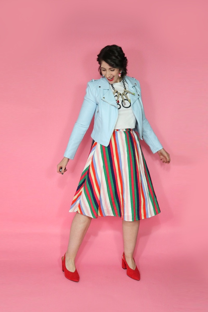 Hannah is wearing a thrifted graphic tee with a blue moto jacket on top, rainbow striped knee length skirt on the bottom, and red high heels