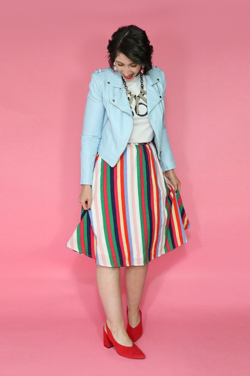 Hannah is wearing a thrifted graphic tee with a blue moto jacket on top, rainbow striped knee length skirt on the bottom, and red high heels