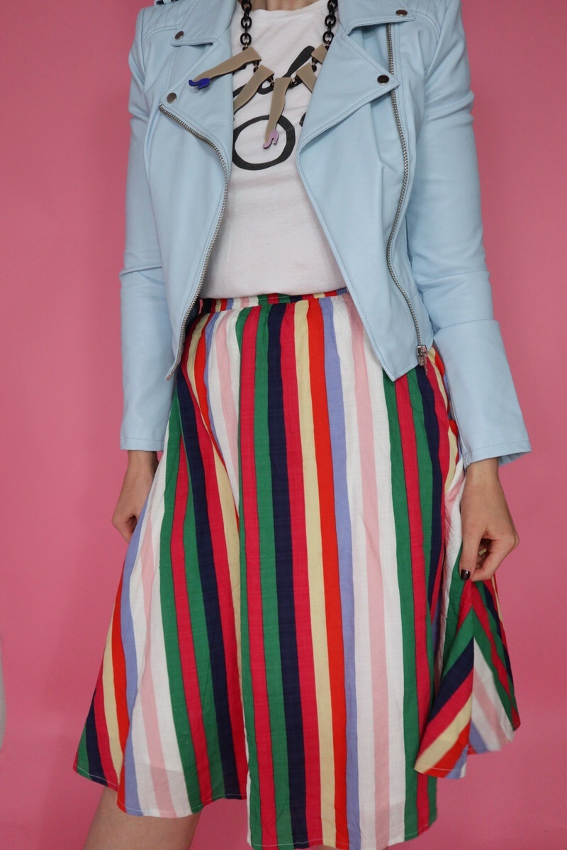 Hannah is wearing a thrifted graphic tee with a blue moto jacket on top, and a rainbow striped knee length skirt on the bottom