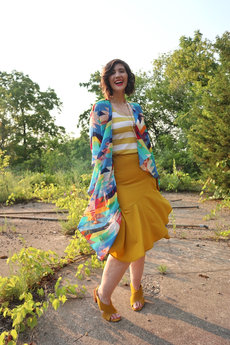 fashion blogger hannah rupp is smiling in an sunny outdoor setting. she's wearing a yellow striped tank top, yellow flouncy skirt, high heels, and a colorful kimono.