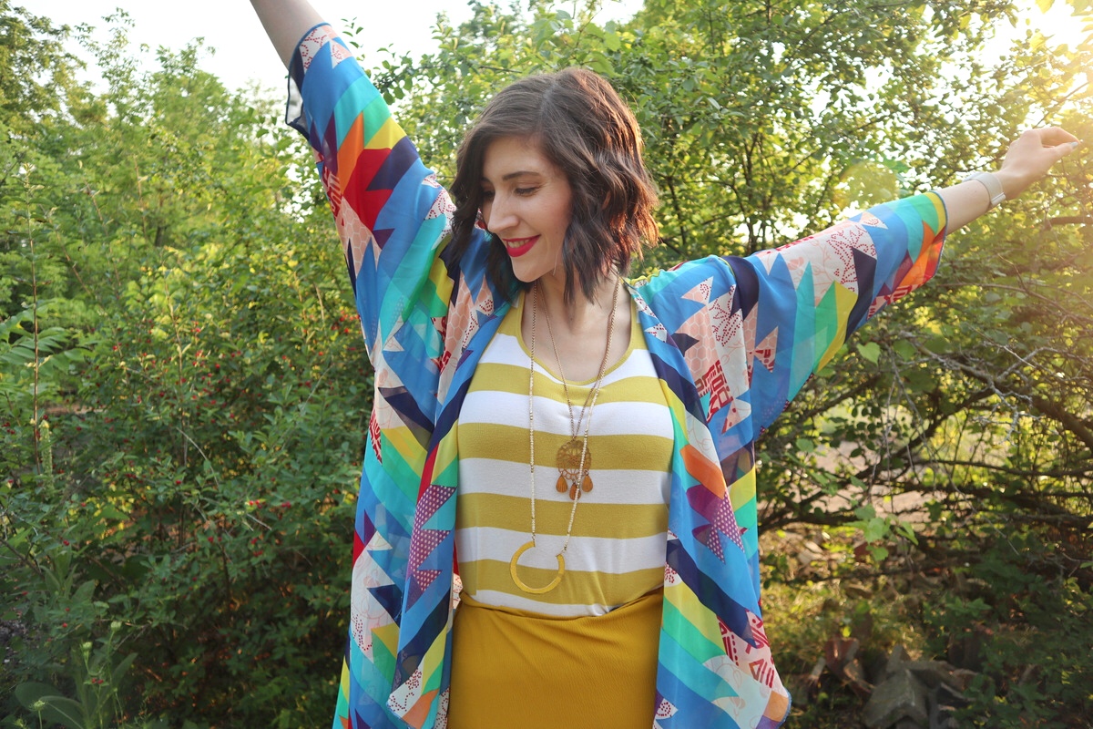 fashion blogger hannah rupp is smiling in an sunny outdoor setting with her arms open wide. she's wearing a yellow striped tank top and a colorful kimono.