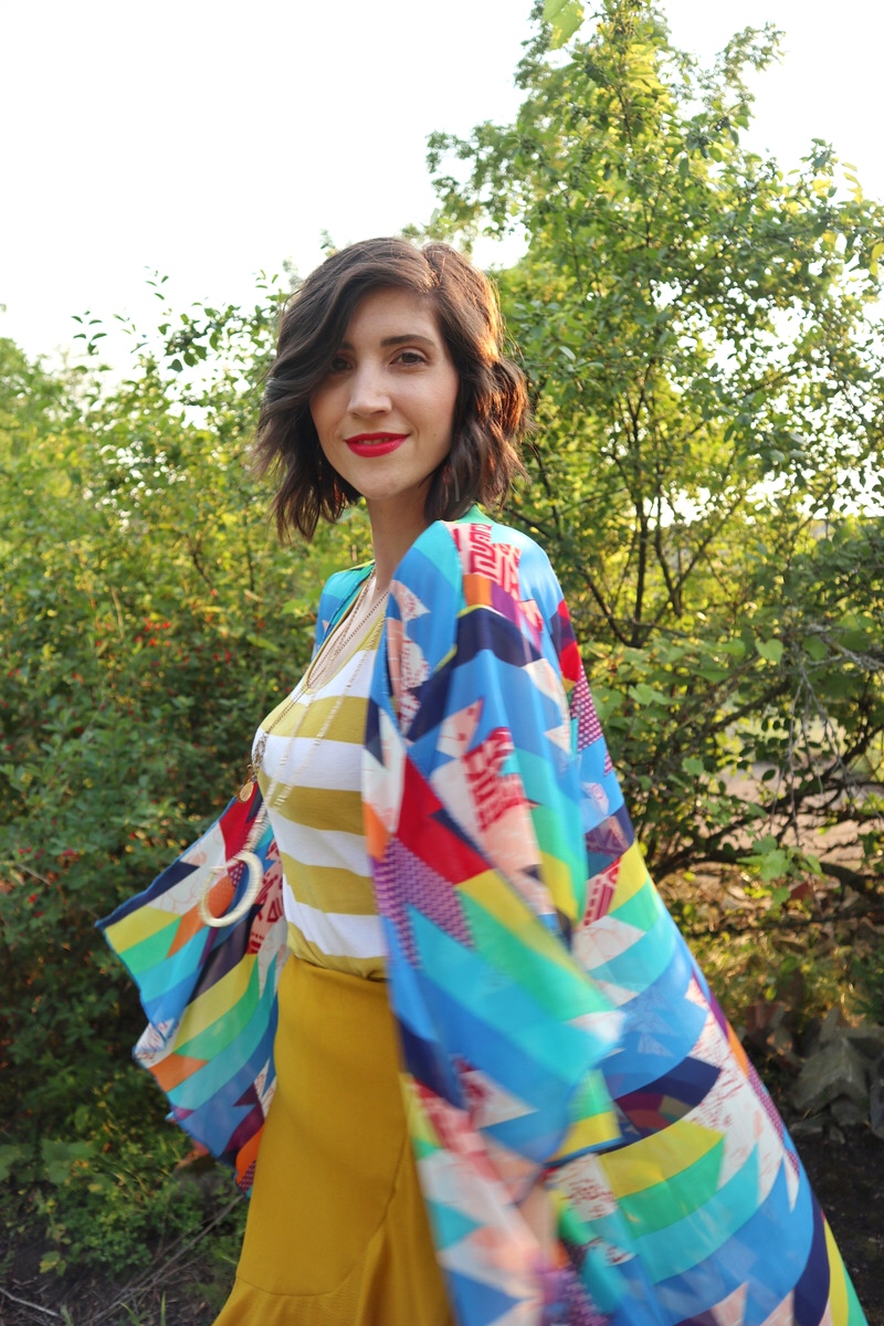 fashion blogger hannah rupp is smiling in an sunny outdoor setting. she's wearing a yellow striped tank top, yellow flouncy skirt, and a colorful kimono.
