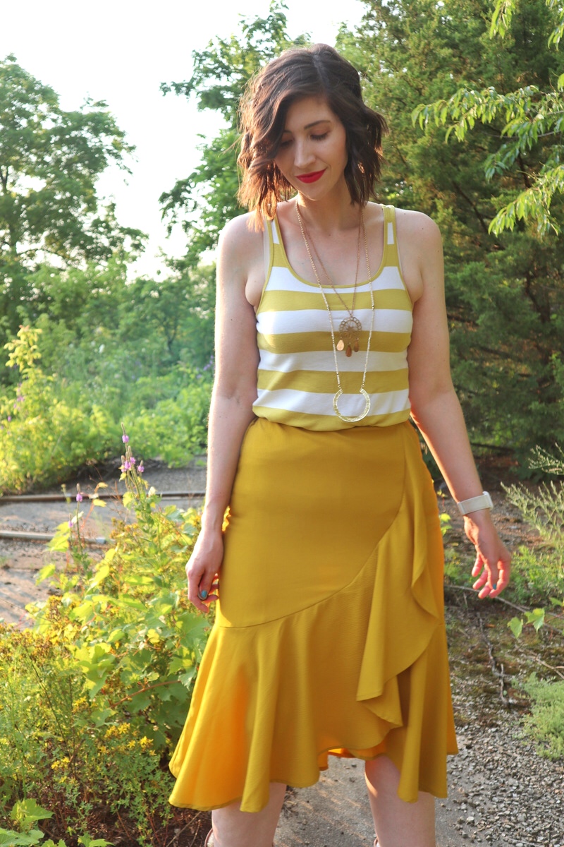 fashion blogger hannah rupp is smiling in an sunny outdoor setting. she's wearing a yellow striped tank top, yellow flouncy skirt, and layered gold necklaces.