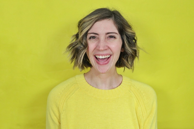 hannah is smiling in front of a solid yellow backdrop while wearing a bright yellow sweater