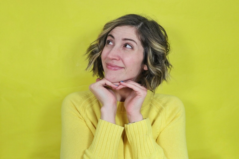 hannah poses with her hands under her chin while gazing upward. she's in front of a yellow backdrop while wearing a bright yellow sweater.