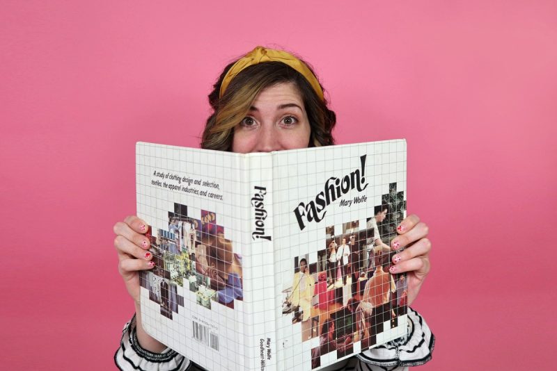 hannah rupp poses in front of a pink background holding a fashion book