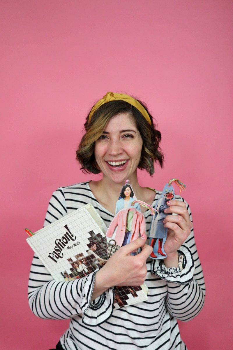 hannah rupp poses in front of a pink background holding a fashion book and bookmarks she made herself