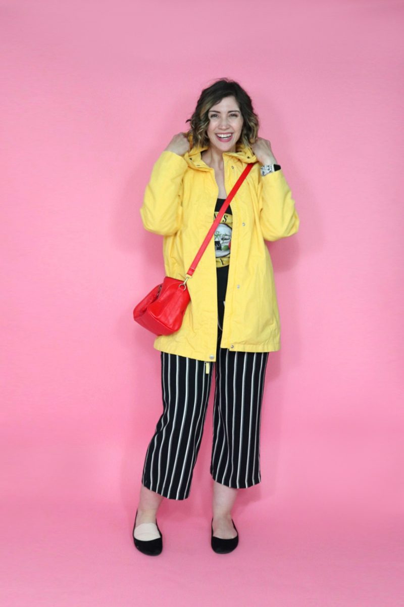 hannah is grinning while popping the collar on a yellow raincoat. underneath are a graphic tee and black striped pants