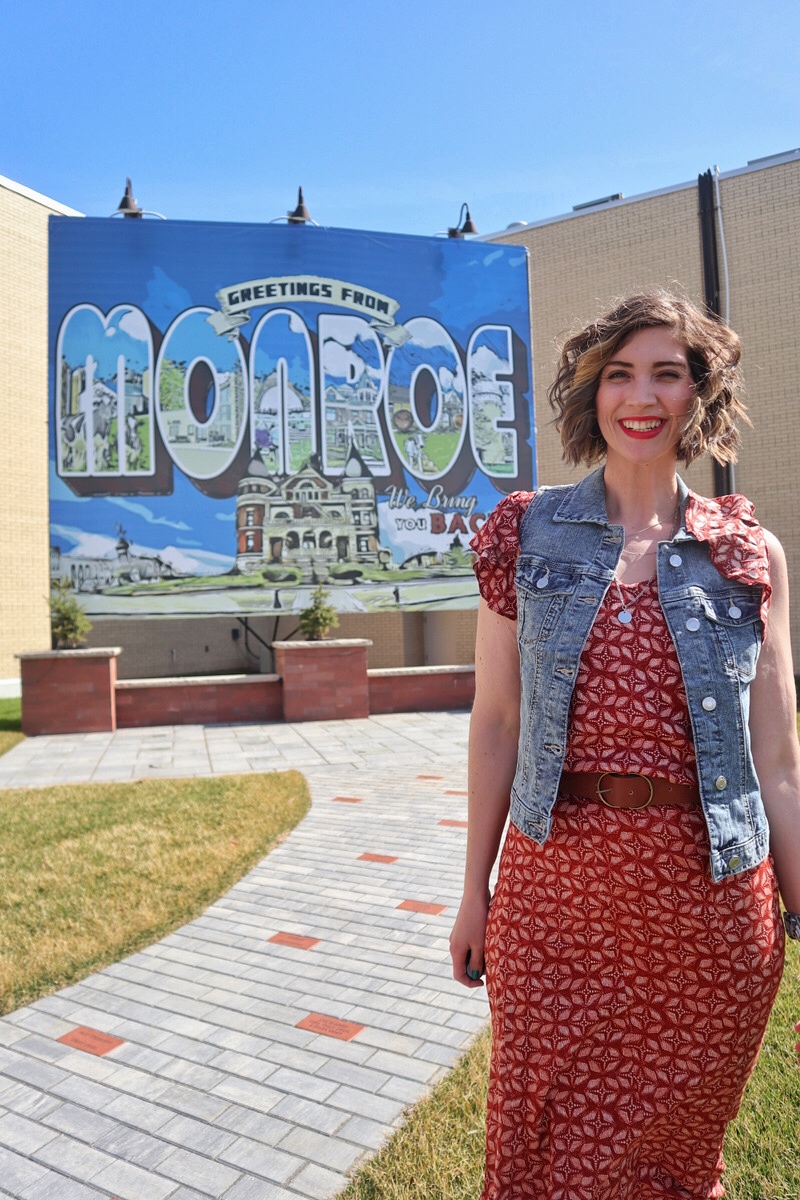 Outfit Photo Dump: What I Wore At Work (In A Clothing Store!) | www.theoutfitrepeater.com hannah is standing in front of a blue billboard that says "greetings from monroe" she's smiling in red lipstick, a red dress, and a denim vest