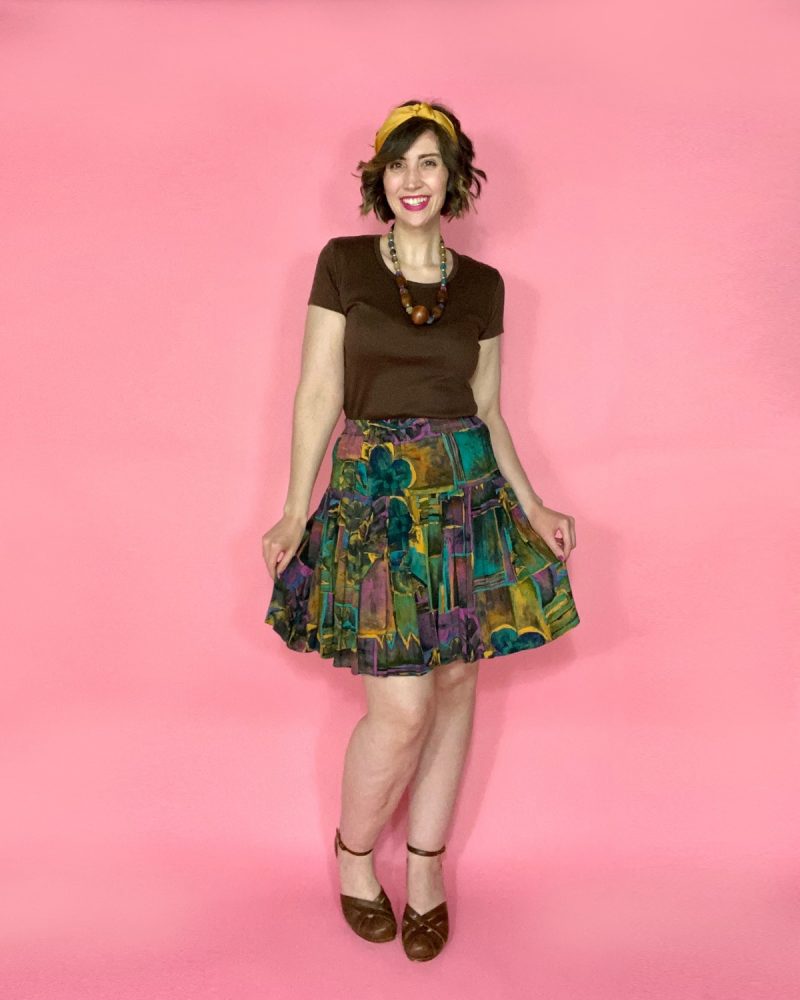 hannah is smiling in front of a pink background wearing a brown t-shirt, colorful skirt, brown high heels, and a mustard yellow headband