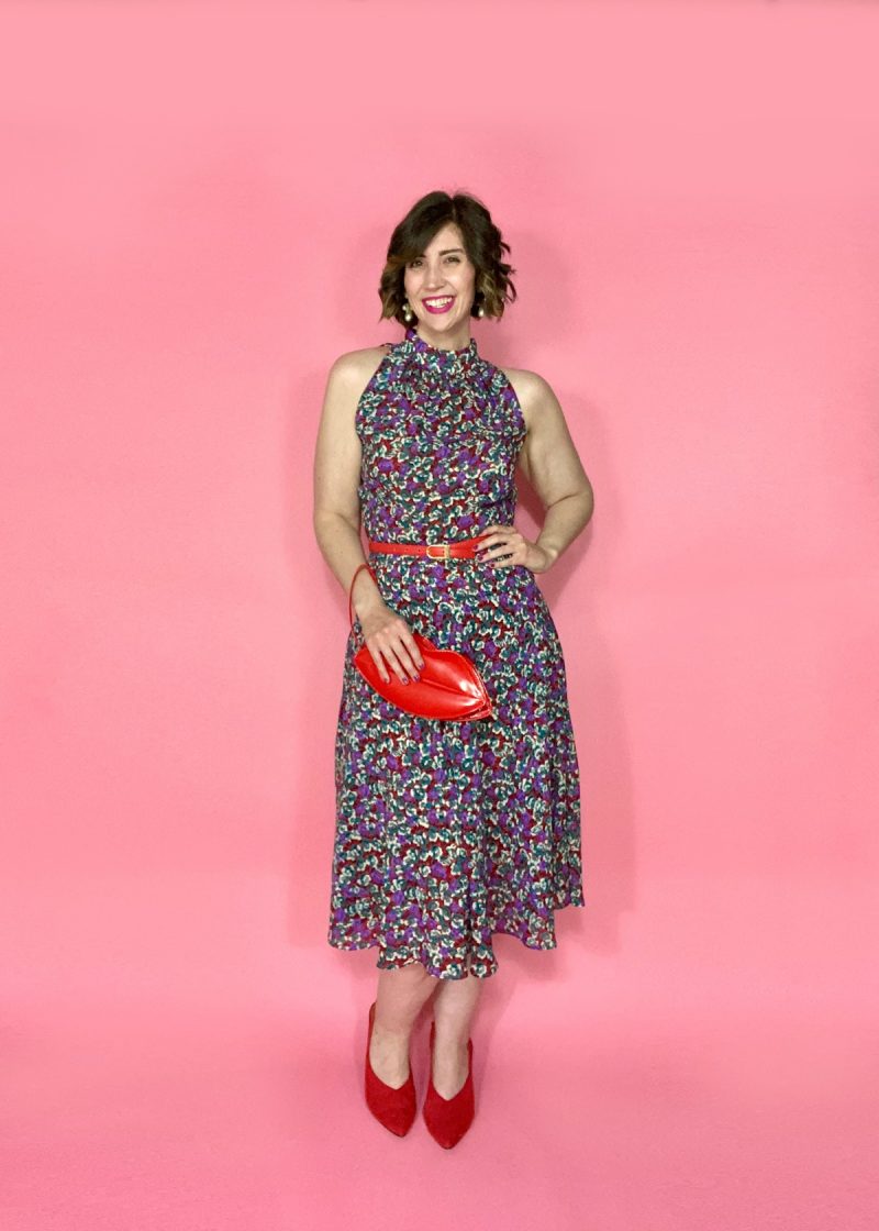 hannah is smiling in front of a pink background wearing a floral sleeveless dress, red belt, red shoes, and a red purse shaped like lips