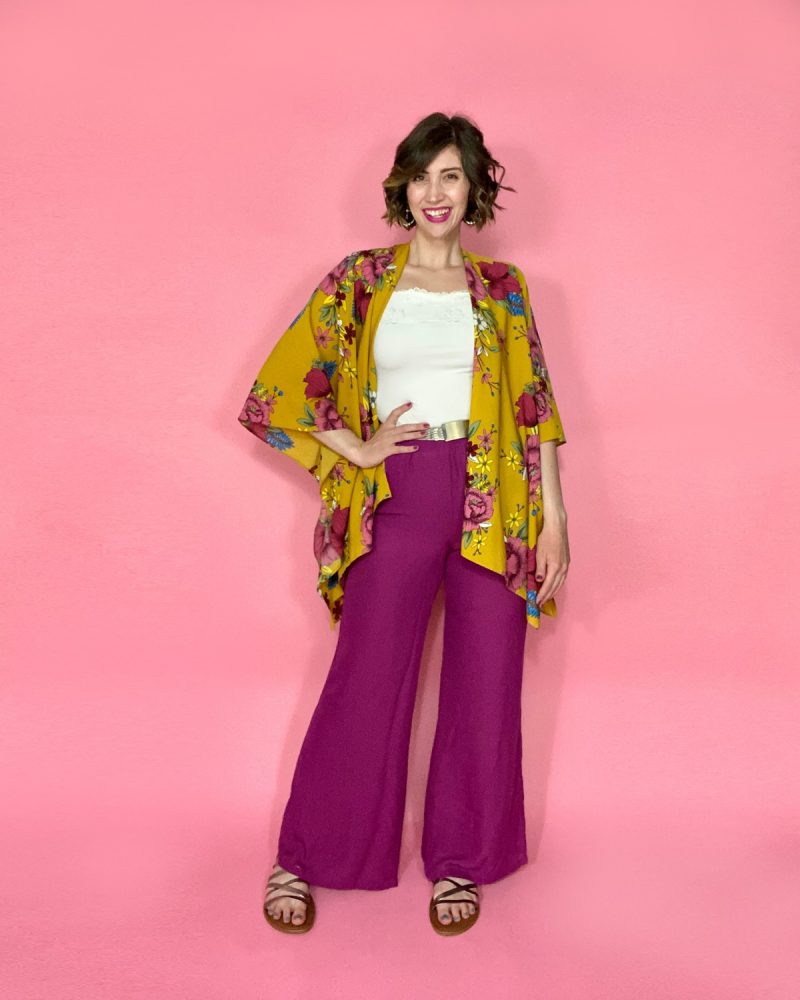 hannah is smiling in front of a pink background. she's wearing a bright yellow kimono with complimenting purple flowers., purple pants, a white tank top, and silver accessories