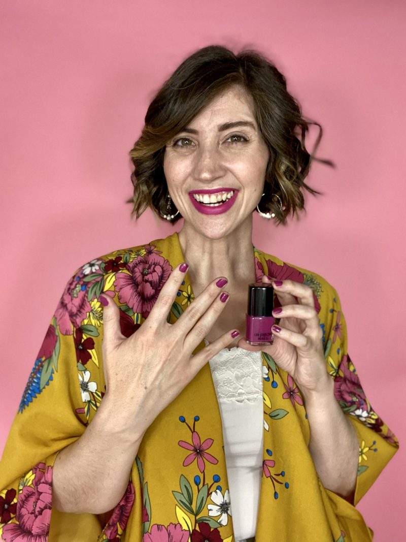 hannah is smiling in front of a pink background and holding a bottle of purple nail polish. she's wearing a bright yellow kimono with complimenting purple flowers.