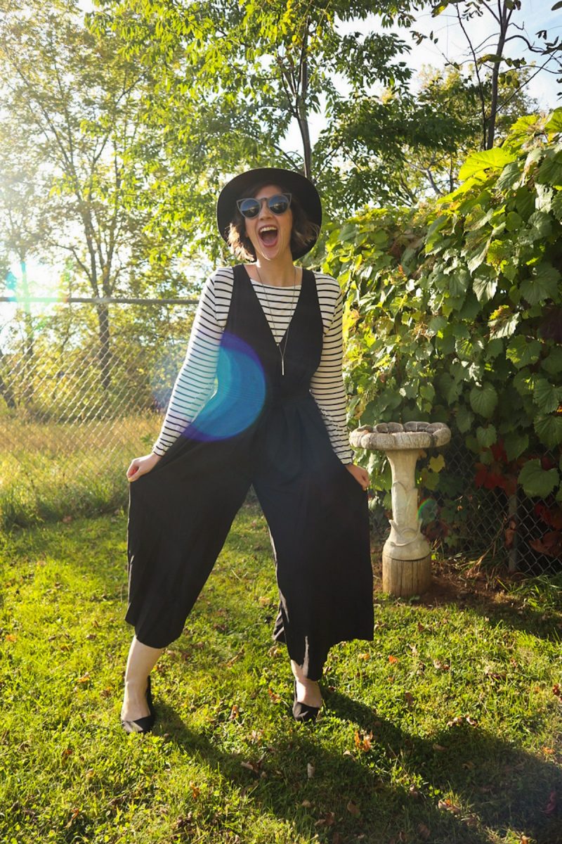 hannah is smiling outside wearing a striped long sleeve shirt, oversized black jumpsuit, and black wide brim fedora