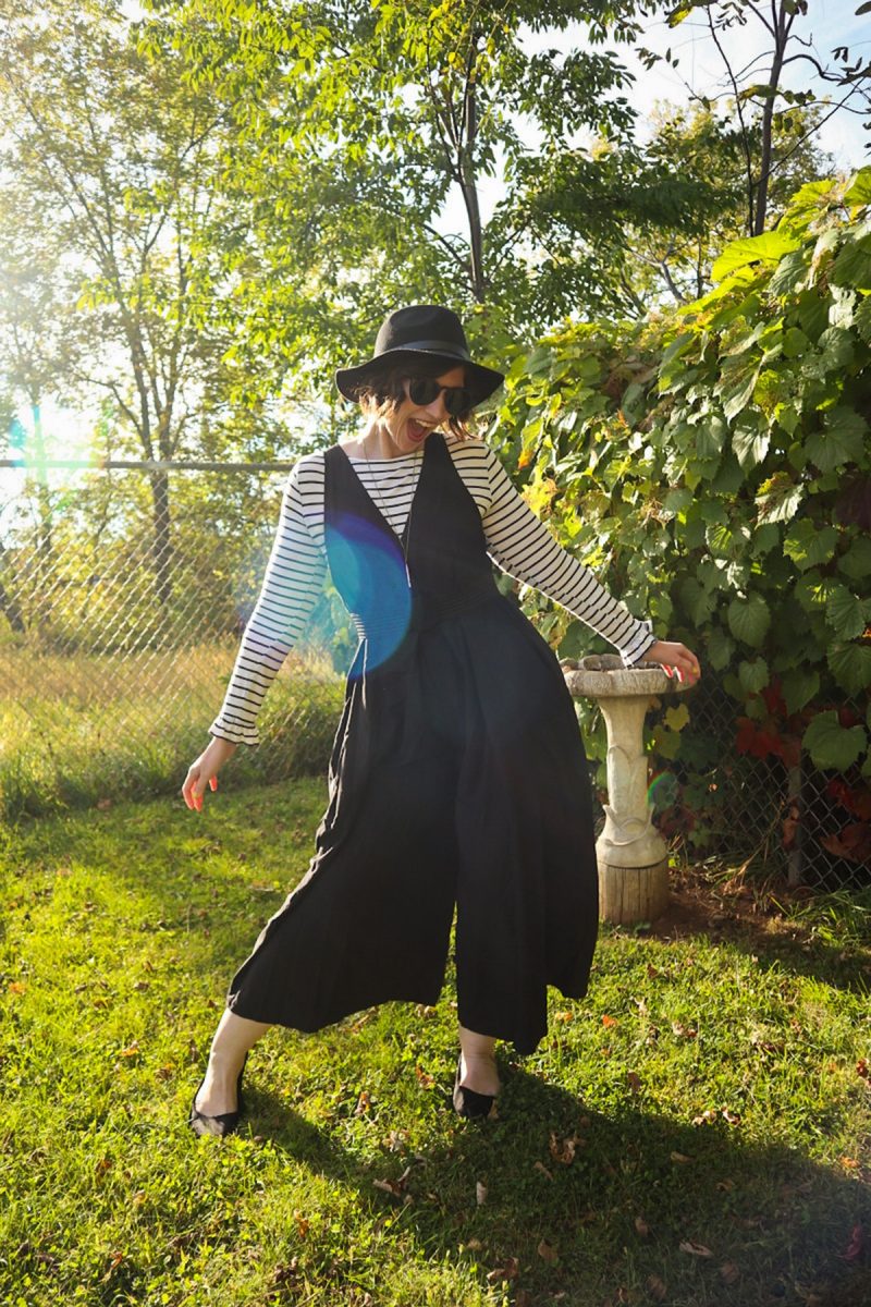 hannah is smiling outside wearing a striped long sleeve shirt, oversized black jumpsuit, and black wide brim fedora