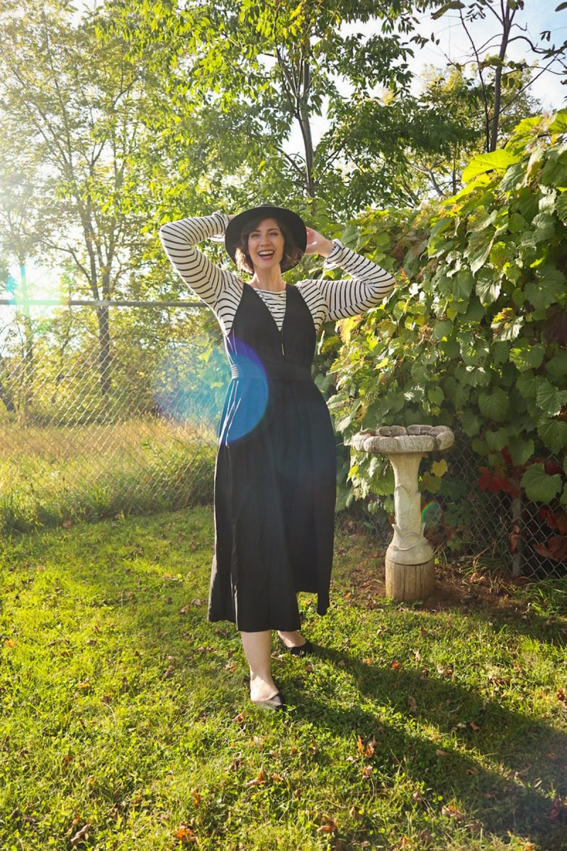 hannah is smiling outside wearing a striped long sleeve shirt, oversized black jumpsuit, and black wide brim fedora