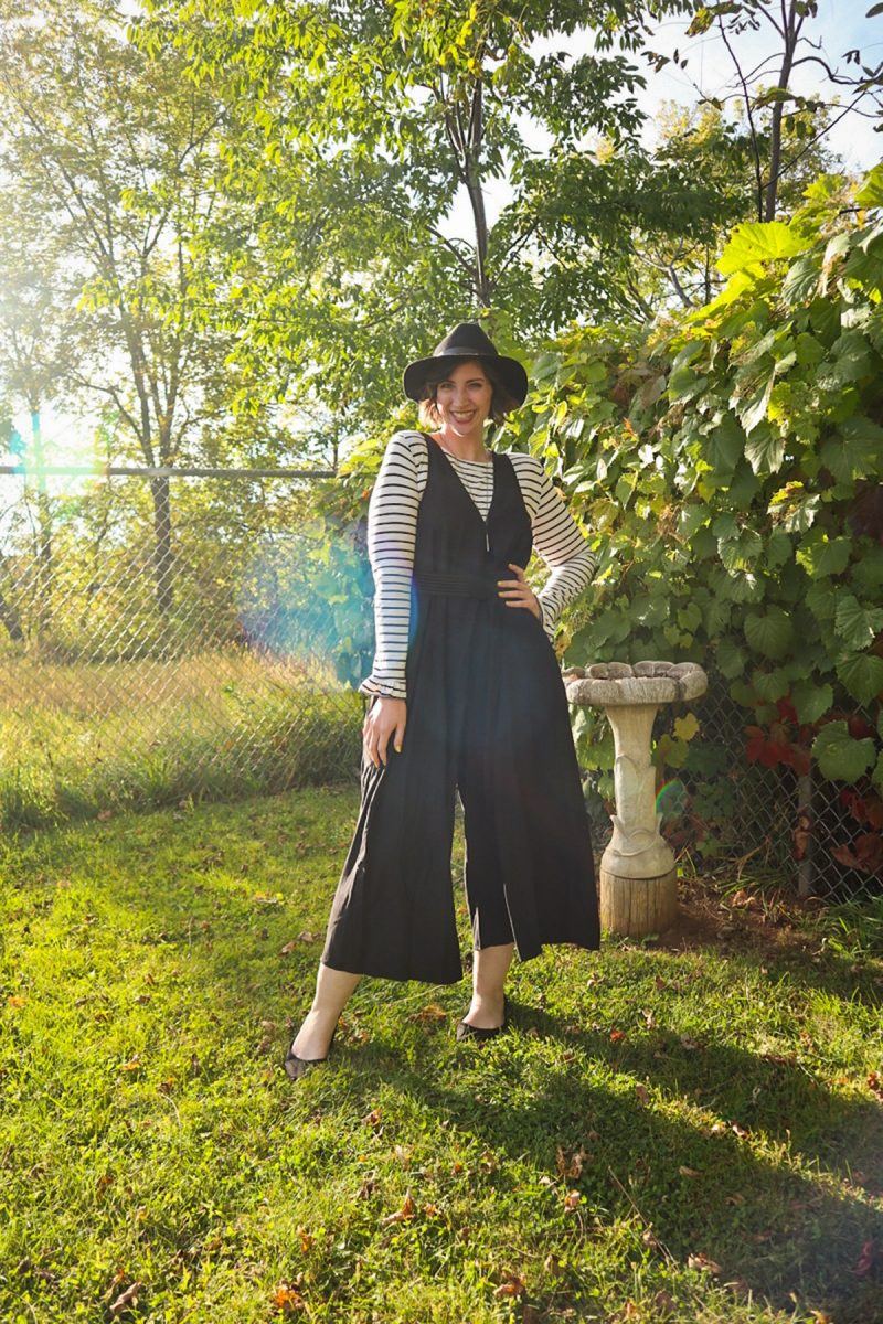 hannah is smiling outside wearing a striped long sleeve shirt, oversized black jumpsuit, and black wide brim fedora