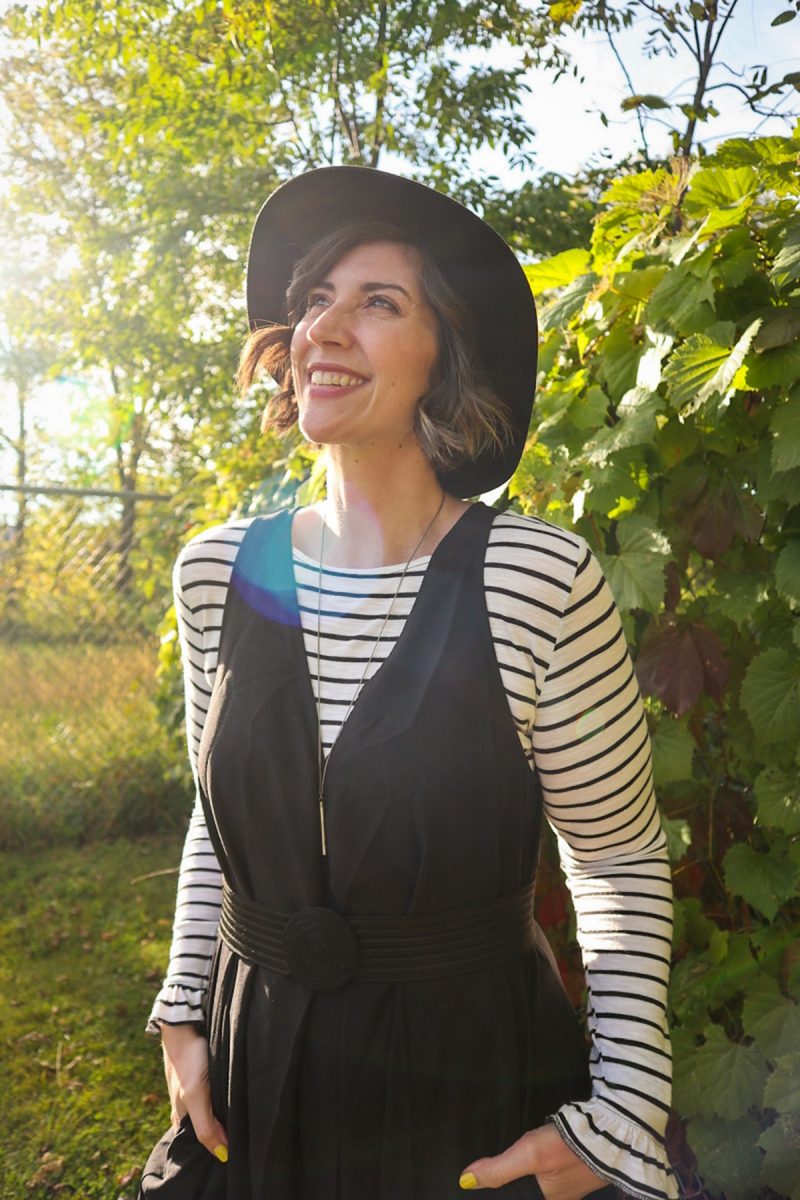 hannah is smiling outside wearing a striped long sleeve shirt, oversized black jumpsuit, and black wide brim fedora