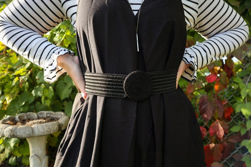 hannah is smiling outside wearing a striped long sleeve shirt, oversized black jumpsuit, and black wide brim fedora