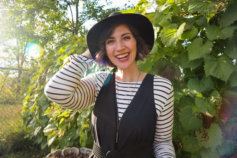 hannah is smiling outside wearing a striped long sleeve shirt, oversized black jumpsuit, and black wide brim fedora