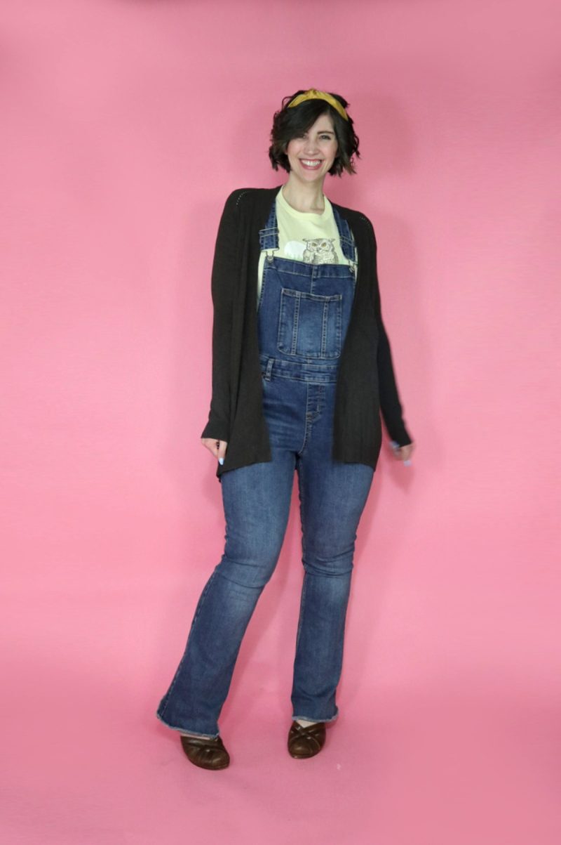 hannah is smiling in front of a pink background. she's wearing a yellow graphic tee, long brown cardigan, and flare denim overalls
