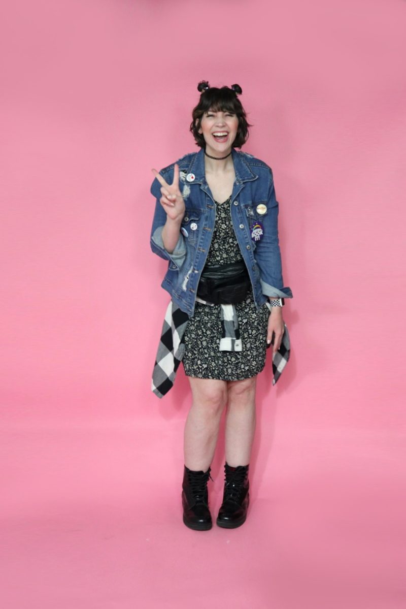 hannah is wearing a 90s themed outfit to drag trivia. her hair is styled in 2 space buns. she's smiling and wearing a black floral dress, distressed denim jacket, a plaid around her waist, a black fanny pack, and black doc marten boots.