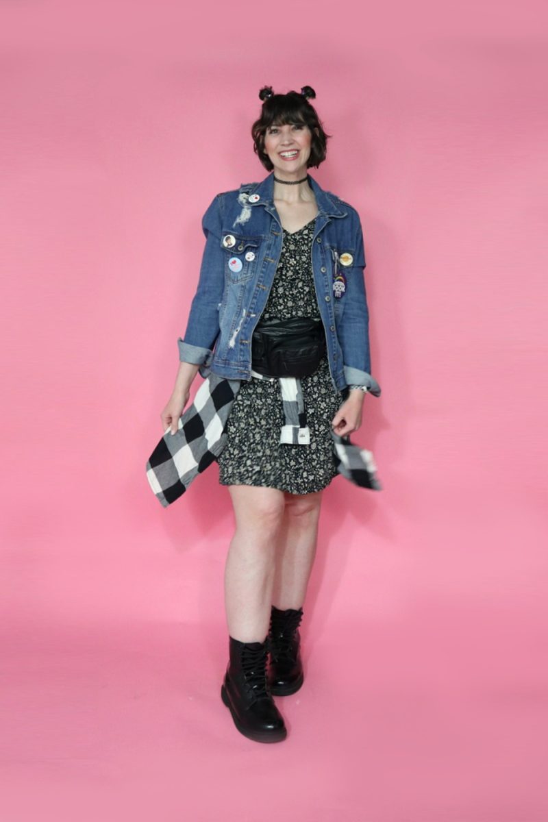 hannah is wearing a 90s themed outfit to drag trivia. her hair is styled in 2 space buns. she's smiling and wearing a black floral dress, distressed denim jacket, a plaid around her waist, a black fanny pack, and black doc marten boots.
