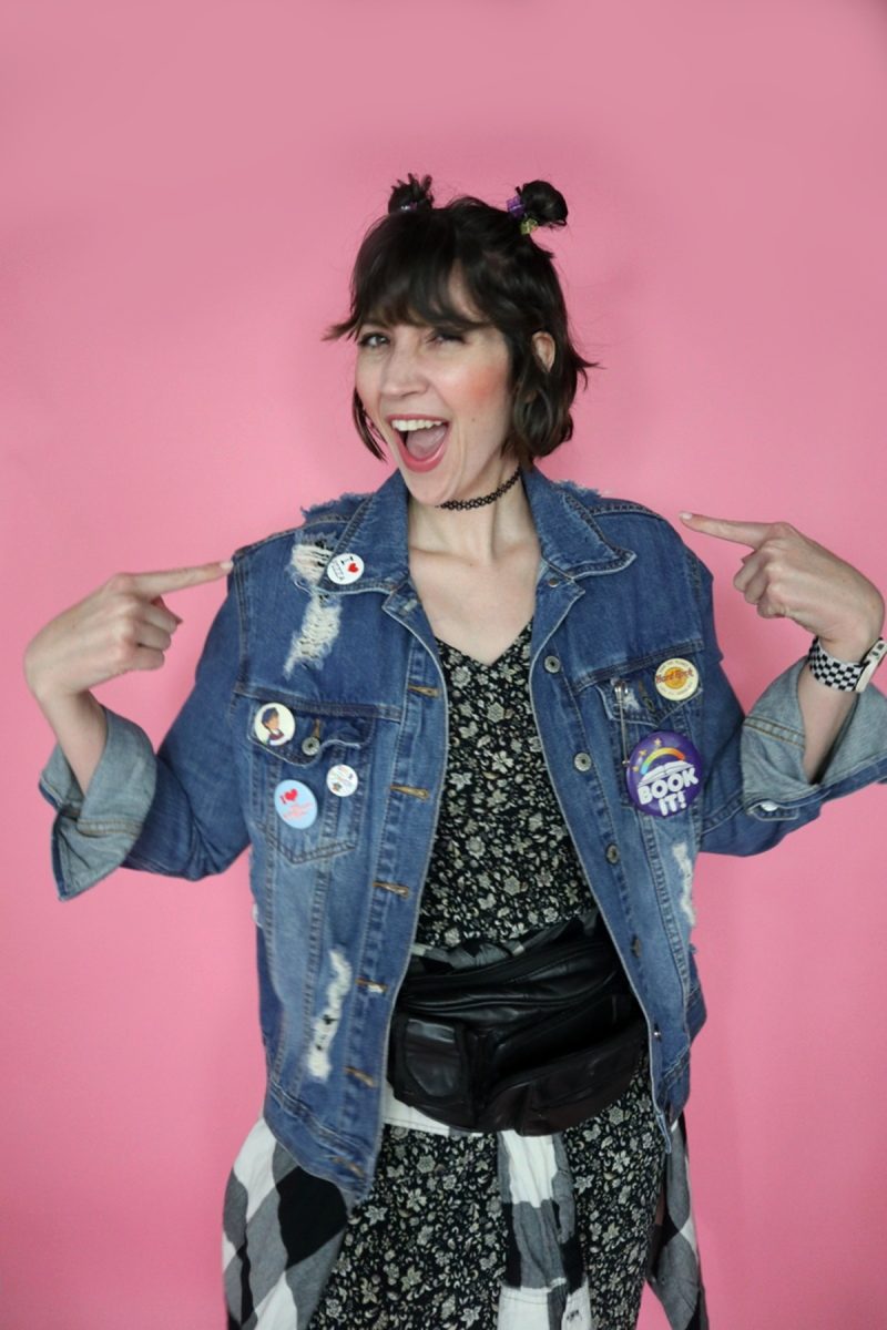 hannah is wearing a 90s themed outfit to drag trivia. her hair is styled in 2 space buns. she's smiling and wearing a black floral dress, distressed denim jacket, a plaid around her waist, and a black fanny pack