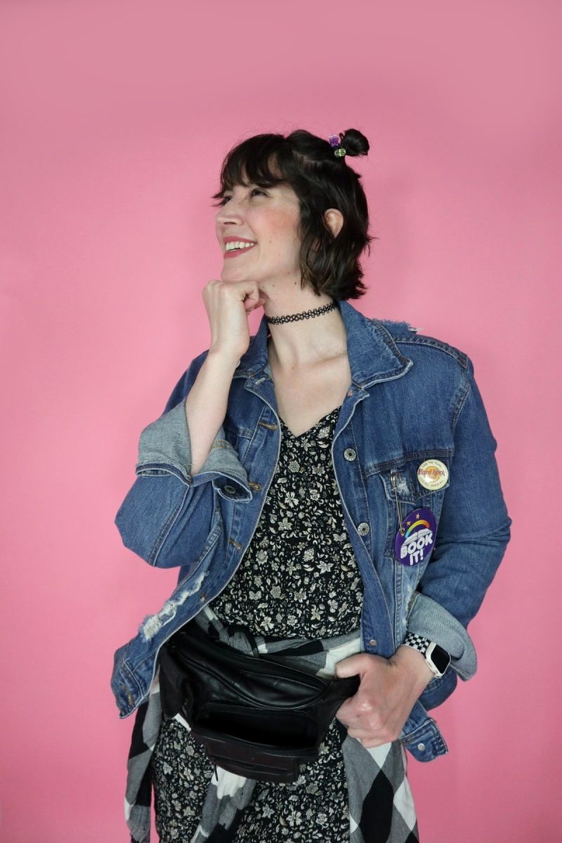 hannah is wearing a 90s themed outfit to drag trivia. her hair is styled in 2 space buns. she's smiling and wearing a black floral dress, distressed denim jacket, a plaid around her waist, and a black fanny pack