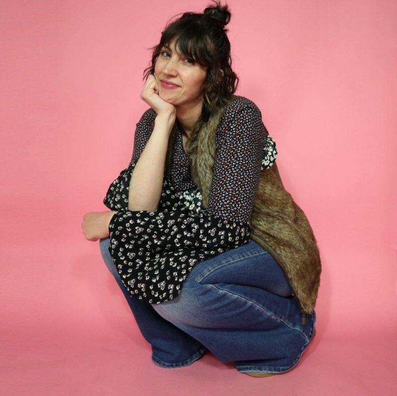 Hannah is white female standing in front of a pink background. Her curly hair is pulled into a bun. She's wearing a floral blouse, brown faux fur vest, light wash flared jeans, and tan booties. 