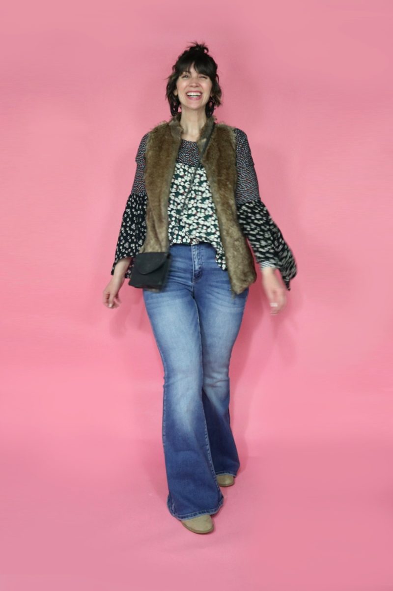 Hannah is white female standing in front of a pink background. Her curly hair is pulled into a bun. She's wearing a floral blouse, brown faux fur vest, light wash flared jeans, and tan booties. 