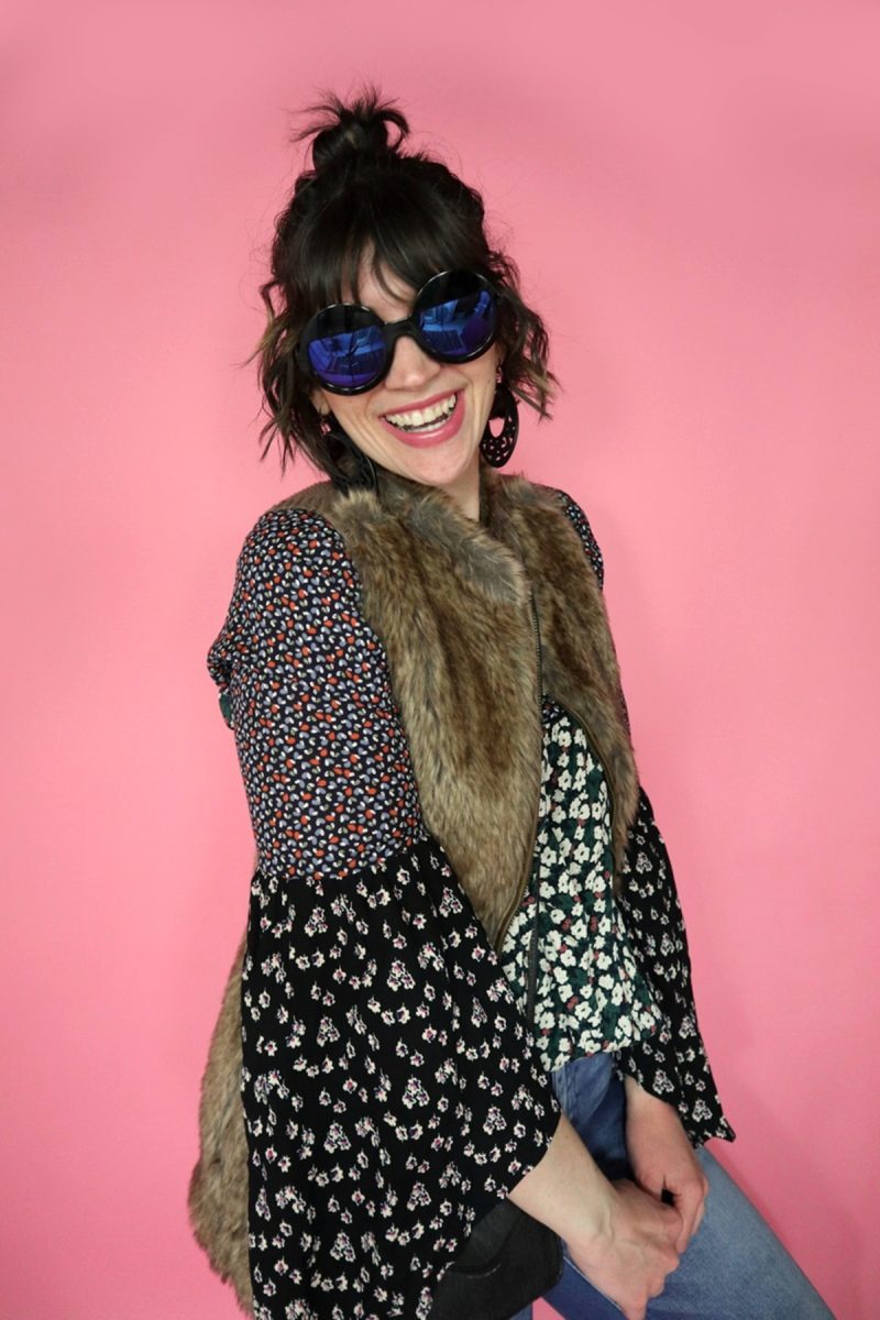 Hannah is white female standing in front of a pink background. Her curly hair is pulled into a bun. She's wearing a floral blouse, brown faux fur vest, and round black sunglasses.
