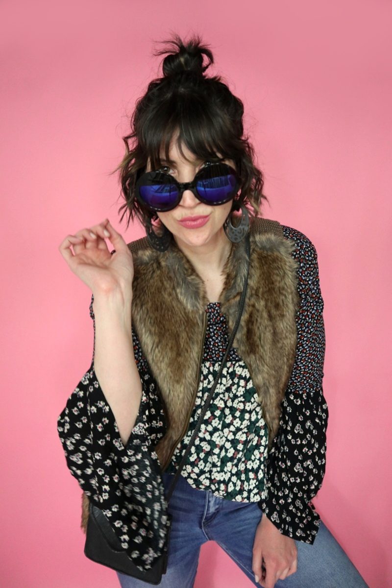 Hannah is white female standing in front of a pink background. Her curly hair is pulled into a bun. She's wearing a floral blouse, brown faux fur vest, and black round sunglasses.