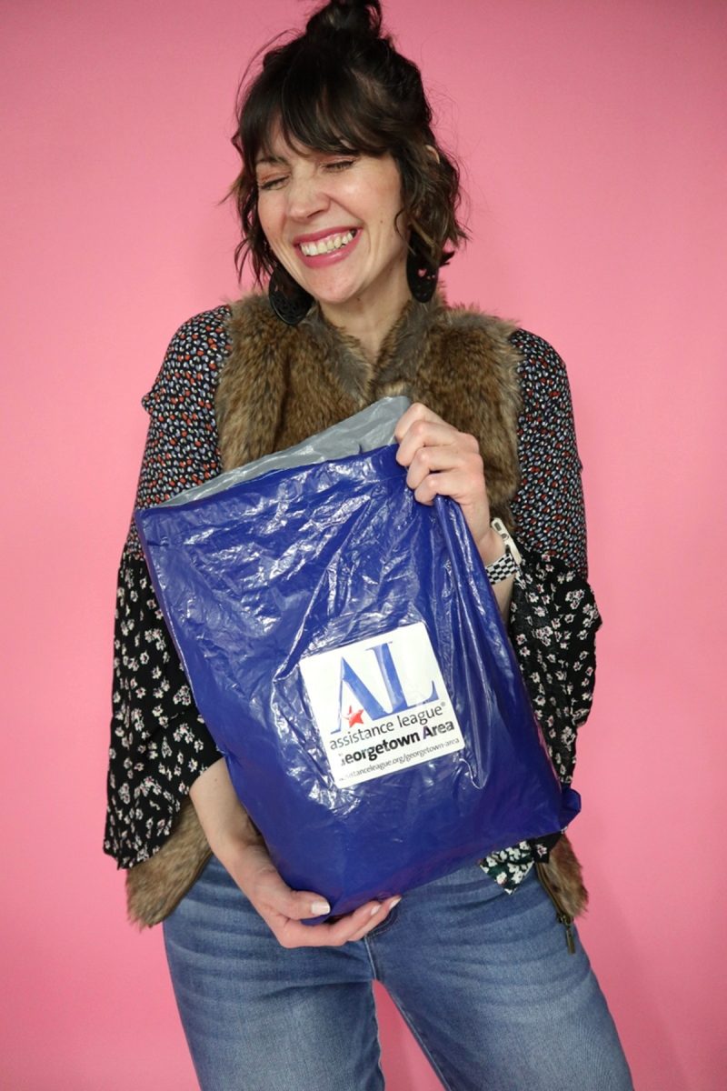 Hannah is standing in front of a pink background. Her curly hair is pulled into a bun. She's smiling in a trendy Spring outfit and holding a blue mailer bag from Assisistance League Online Thrift Shop