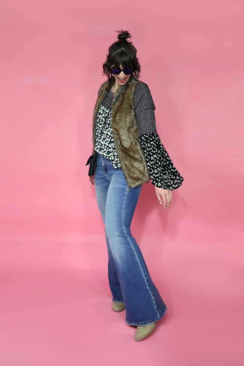 Hannah is white female standing in front of a pink background. Her curly hair is pulled into a bun. She's wearing a floral blouse, brown faux fur vest, black round sunglasses, light wash flared jeans, and tan booties. 