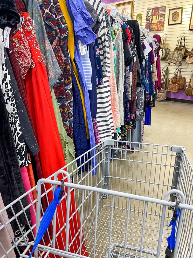 a gray metal cart in a thrift store aisle, facing a rack of colorful clothes hanging up.