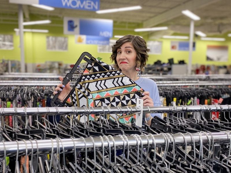 Hannah is standing in a thrift store, in front of a clothing rack. She's holding up a brightly colored beaded mini skirt and shrugging.