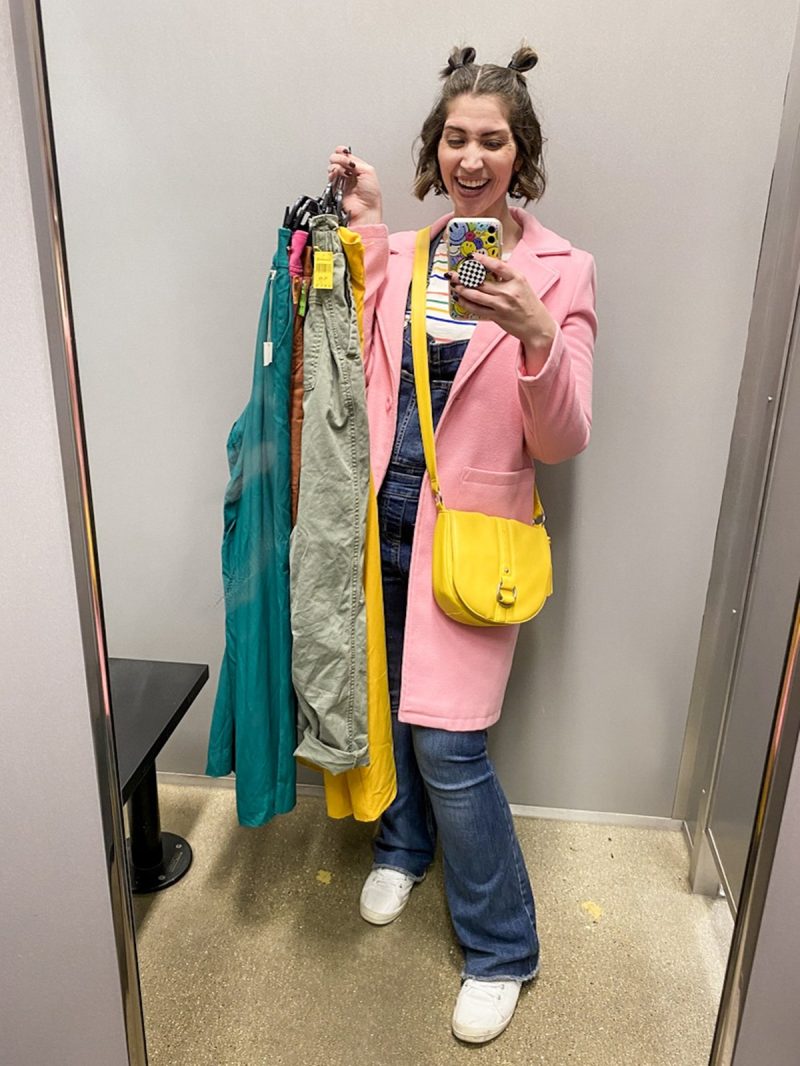 Hannah is smiling a thrift store fitting room holding a pile of colorful clothes. she's wearing a long pink jacket, denim overalls, and a yellow purse.