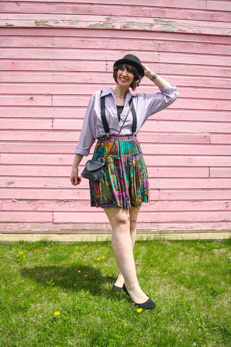 hannah is standing in front of a pink wall. she's wearing a black hat, purple striped button, black suspenders, colorful vintage skirt, and black flat shoes.