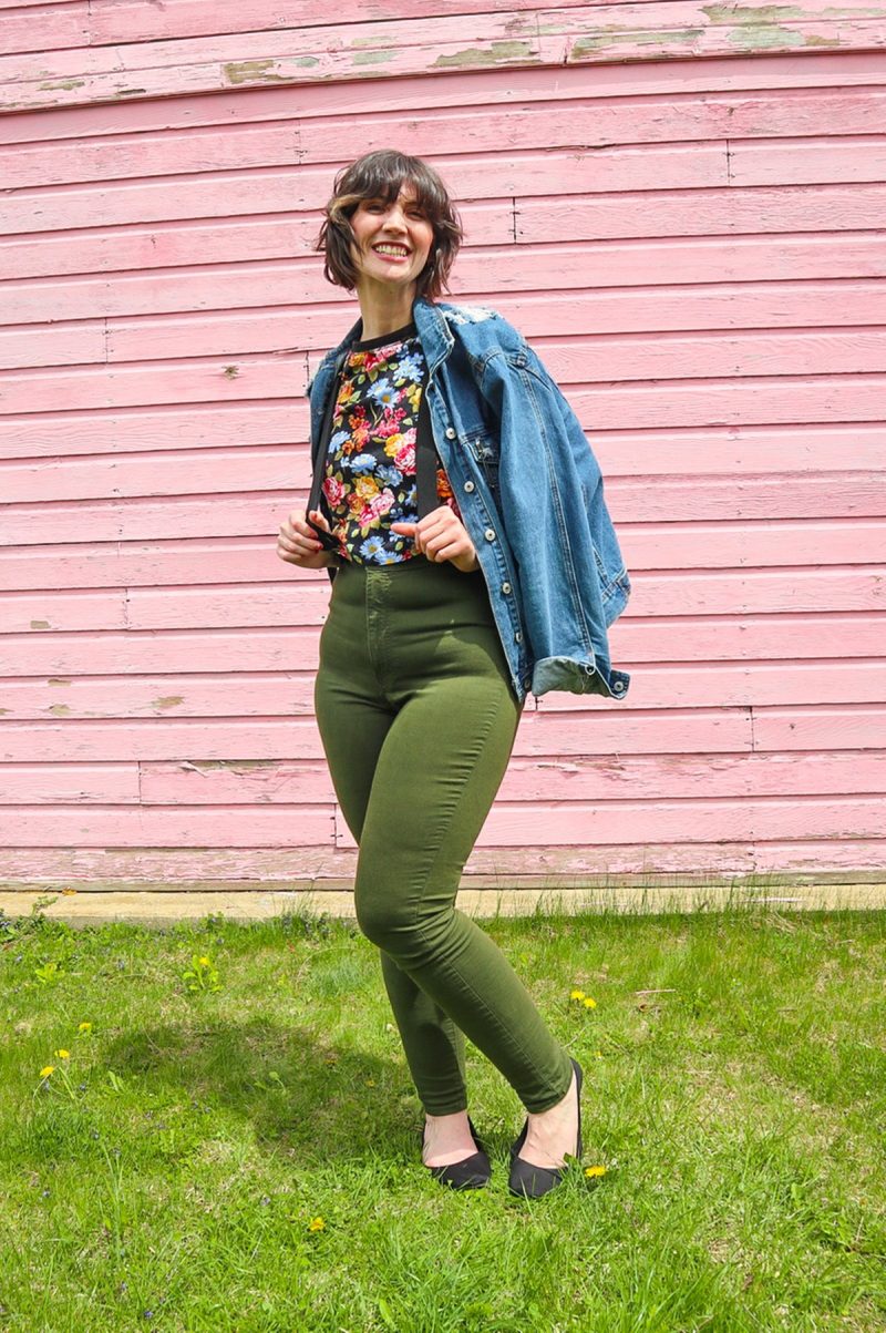 How To Wear Suspenders For Women: 3 Feminine Outfit Ideas | theoutfitrepeater.com hannah is standing in front of a pink wall. she's wearing a floral crop top, denim jacket, black suspenders, olive green skinny pants, and a black flat shoes.